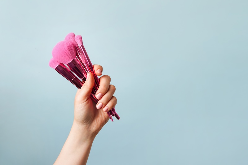 Eine Hand Hält Einen Satz Rosa MakeUpPinsel Vor Einem Hellblauen Hintergrund. Die Szene Vermittelt Ein Gefühl Von Eleganz Und Schönheit Und Hebt Kosmetische Werkzeuge Hervor.