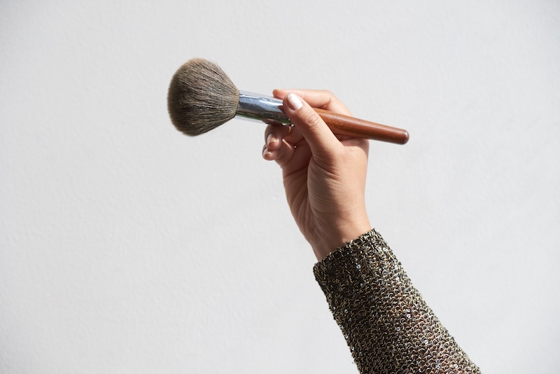 Eine Hand Hält Einen MakeUpPinsel Mit Holzgriff Vor Hellgrauem Hintergrund. Der Ärmel Ist Aufwendig Gemustert Und Verleiht Ihm Eine Elegante Note.