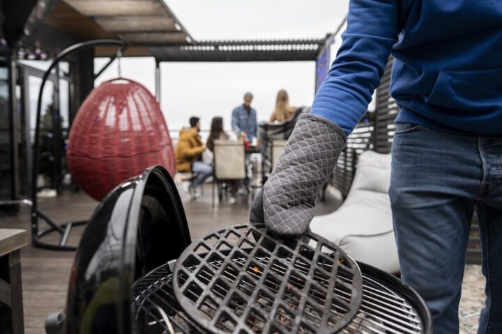 A person in a blue sweatshirt lifts a grill grate with oven mitts on a patio. In the background, four people socialize near a red egg chair.