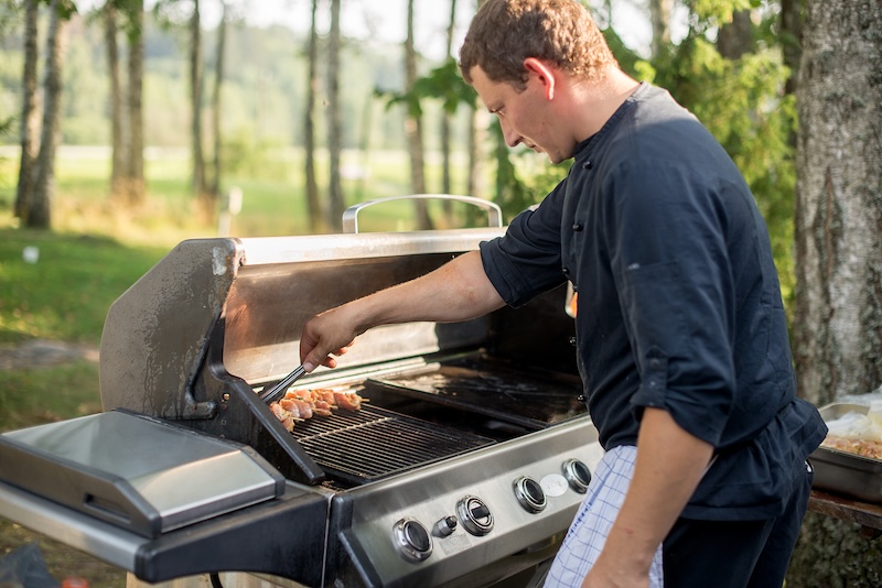 Man Grilling Skewers On An Outdoor Barbecue, Dressed In A Dark Chef's Uniform. Sunlight Filters Through Nearby Trees, Enhancing The Serene Setting.