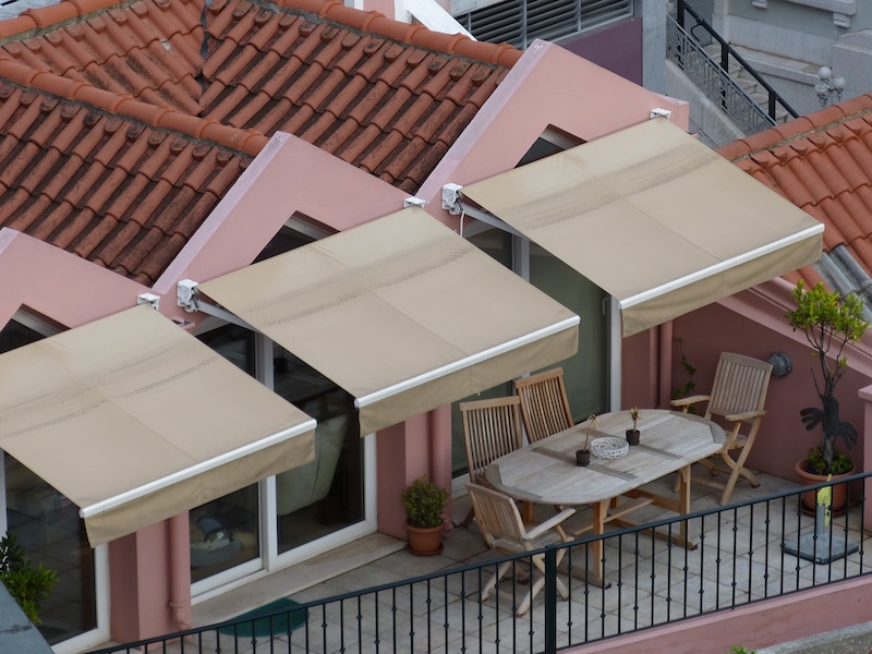 Die Terrasse Mit Pfirsichfarbenen Wänden Und Rotem Ziegeldach Verfügt Über Drei Beige Markisen, Hölzerne Esszimmermöbel Und Topfpflanzen Und Schafft So Einen Gemütlichen, Einladenden Außenbereich.