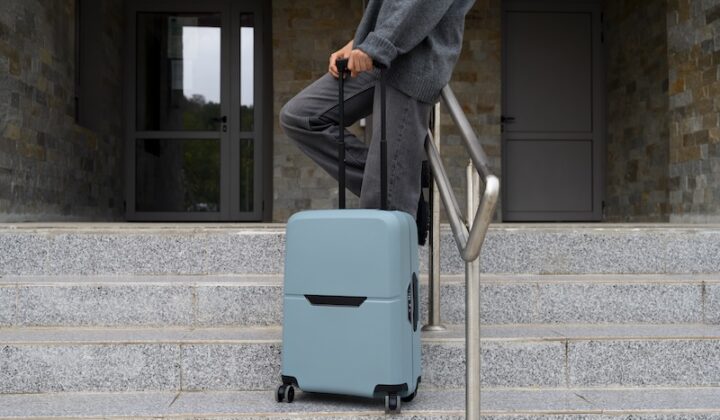 Person in gray clothing standing on steps, holding the handle of a large light blue suitcase. The scene suggests travel or departure.