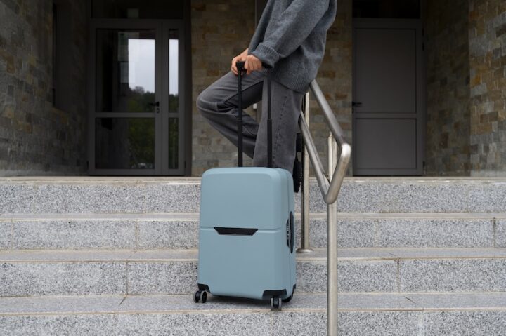 Person in gray clothing standing on steps, holding the handle of a large light blue suitcase. The scene suggests travel or departure.