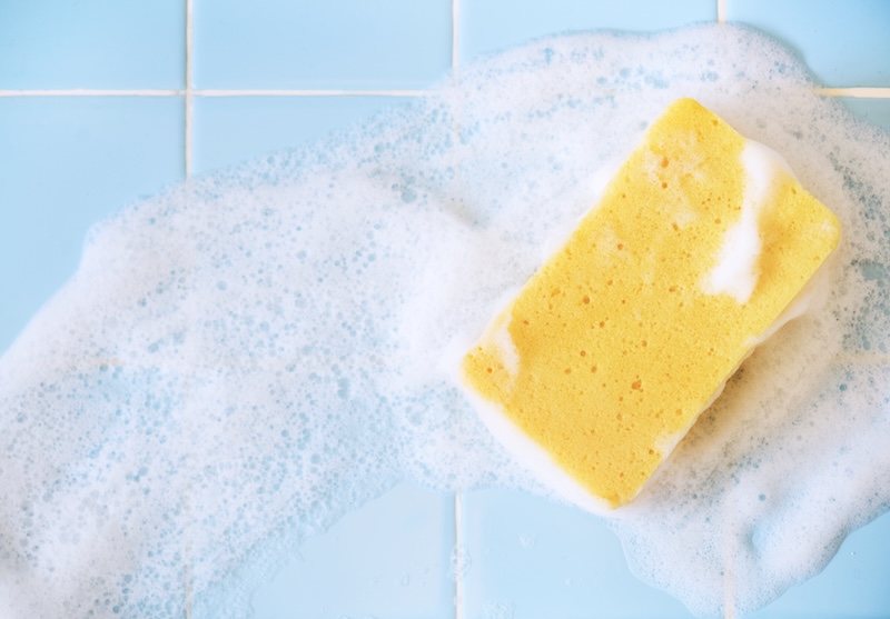 Ein Gelber Schwamm Mit Weißem Schaumstoff Ruht Auf Hellblauer Fliesenoberfläche. Die Szene Vermittelt Ein Frisches, Sauberes Gefühl Und Hebt Das Reinigen Oder Waschen Hervor.
