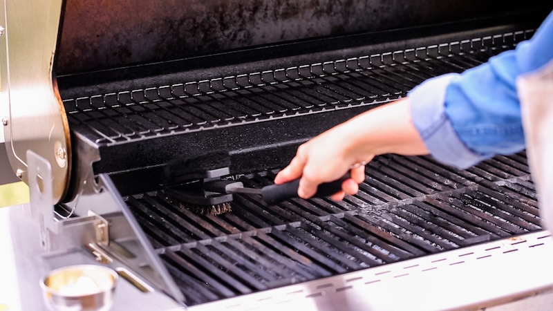 A Person In A Blue Shirt Cleans A Grill With A Brush, Focusing On The Blackened Grates. The Image Conveys A Sense Of Cleanliness And Preparation.