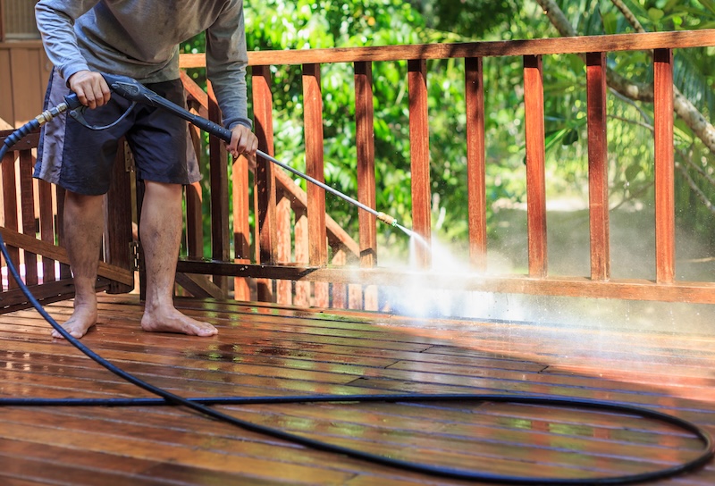 A Person In Shorts Pressure Washes A Wooden Deck. Sunlight Filters Through Trees In The Background, Creating A Fresh And Vibrant Outdoor Setting.