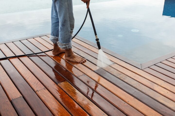 A person in rolled-up jeans is pressure washing a wooden deck by a calm pool. The scene conveys a sense of cleanliness and tranquility.