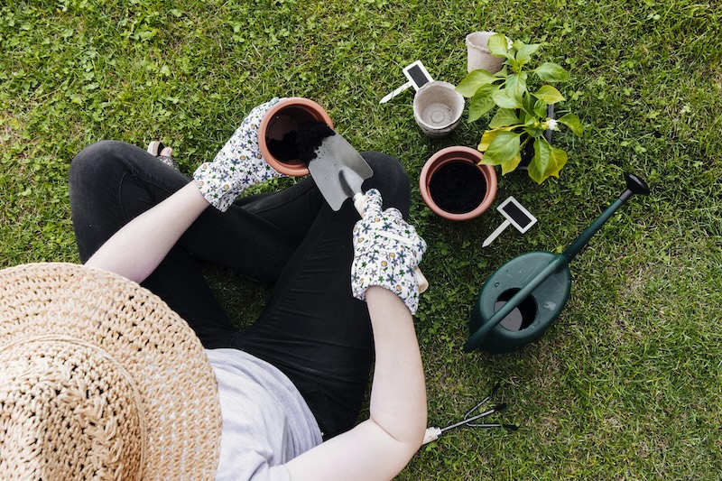Eine Person Mit Strohhut Und Blumenhandschuhen Sitzt Auf Gras Und Pflanzt Pflanzen Ein. In Der Nähe Befinden Sich Setzlinge, Töpfe, Eine Gießkanne Und Gartengeräte, Die Gelassenheit Vermitteln.