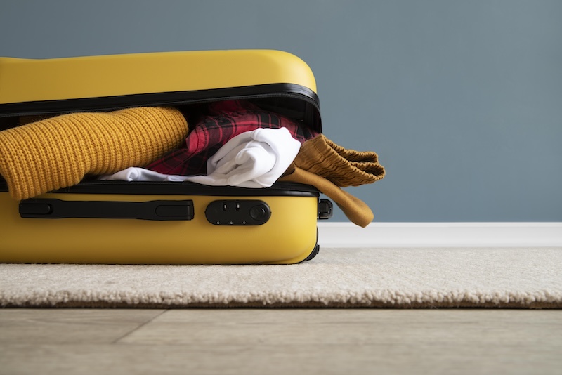 A Yellow Suitcase Is Overfilled With Clothes, Including A Mustard Sweater, A Red Plaid Shirt, And A White Top, Spilling Out Onto A Beige Carpet.