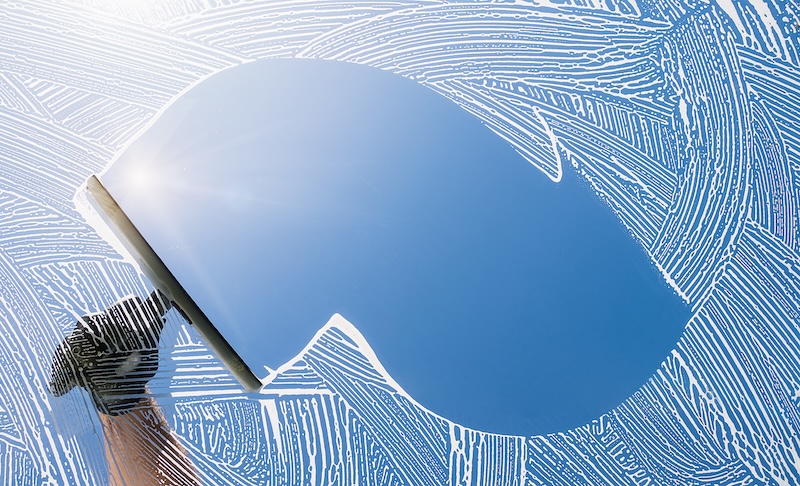 A Hand Uses A Squeegee To Clean A Window, Revealing A Clear Blue Sky And Sunburst Through Swirling Soap Streaks. The Scene Feels Fresh And Crisp.