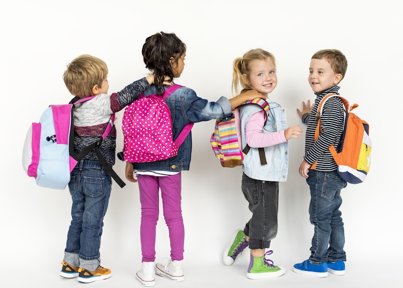 Vier Kinder Stehen In Einer Reihe, Jedes Trägt Bunte Rucksäcke Und Blickt Zur Seite. Sie Wirken Fröhlich Und Verspielt, Mit Einem Schlichten Weißen Hintergrund.