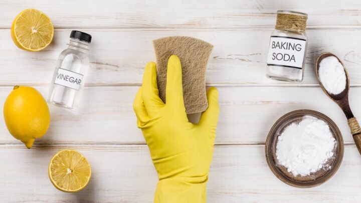 A person wearing a yellow glove cleans a sponge with lemon and various cleaning supplies on a countertop.