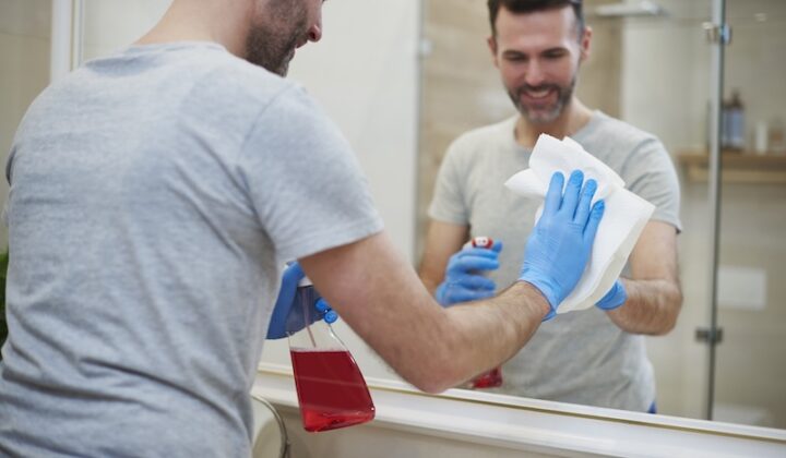 Mann in grauem Hemd und blauen Handschuhen wäscht sich die Hände vor einem Spiegel.