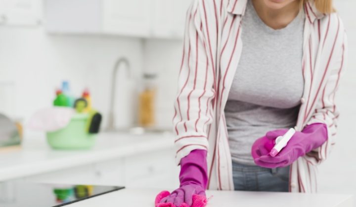 A person wearing pink gloves and a striped shirt is cleaning a kitchen counter with a spray bottle and cloth. The setting appears bright and tidy.