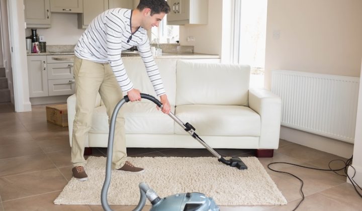 Man in his twenties cleaning area rug with canister vacuum in living room, copy space. Modern, home, lifestyle, cleaning, interior, minimalism, productivity