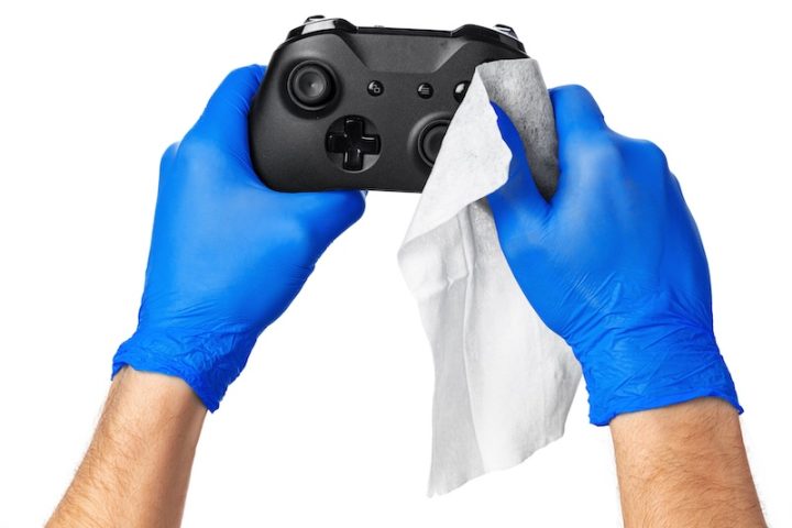 Hands in blue gloves clean a black game controller with a white cloth, set against a plain white background, conveying cleanliness and care.