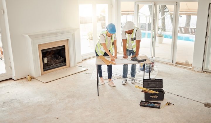 Two construction workers in hard hats and vests review blueprints on a table in a bright, unfinished room with a fireplace and large windows.