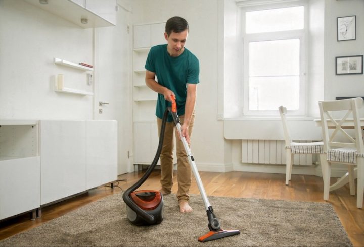 A man in a green shirt vacuums a carpet in a bright, minimalist room. The space has white walls, wooden floors, and simple furniture, conveying cleanliness.