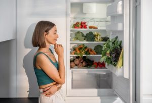 A woman with a thoughtful expression stands beside an open fridge filled with fresh vegetables like carrots, broccoli, and greens, in a brightly lit kitchen.