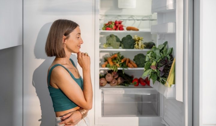A woman with a thoughtful expression stands beside an open fridge filled with fresh vegetables like carrots, broccoli, and greens, in a brightly lit kitchen.