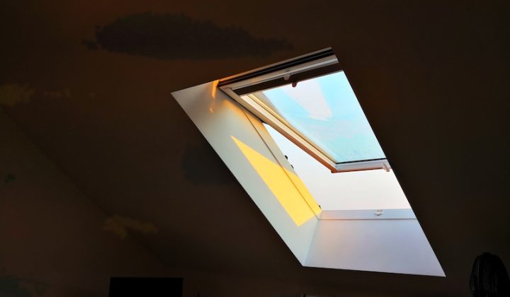 Sunlight streams through an open skylight, casting a warm glow on a dim attic room. A small TV and radiator are partially visible in the shadows.