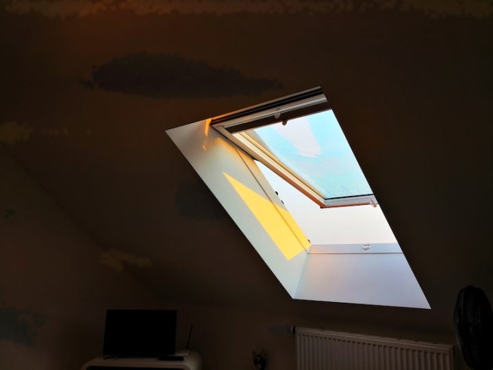 Sunlight streams through an open skylight, casting a warm glow on a dim attic room. A small TV and radiator are partially visible in the shadows.