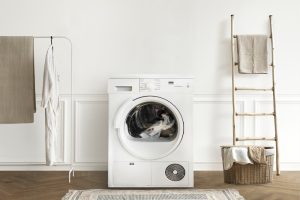 A white washing machine in a laundry room, with detergent bottles and a potted plant on top. Towels hang nearby, and an iron rests on an ironing board.