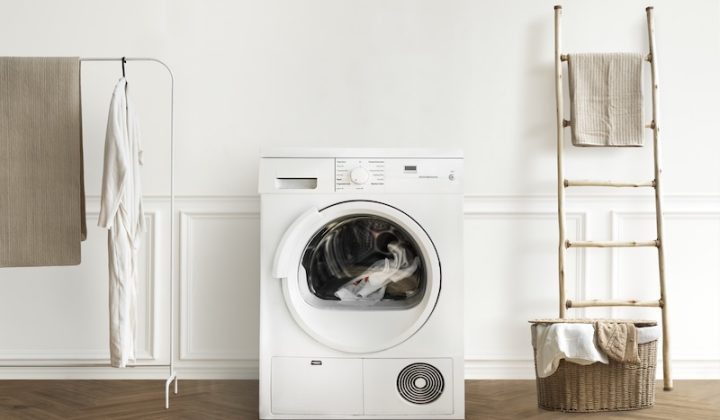 A white washing machine in a laundry room, with detergent bottles and a potted plant on top. Towels hang nearby, and an iron rests on an ironing board.
