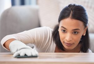 A focused woman cleans a wooden surface with a cloth, wearing a white shirt and gloves. Her expression shows concentration and attention to detail.