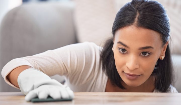 A focused woman cleans a wooden surface with a cloth, wearing a white shirt and gloves. Her expression shows concentration and attention to detail.