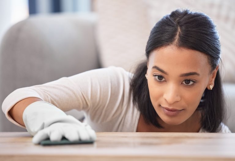 A focused woman cleans a wooden surface with a cloth, wearing a white shirt and gloves. Her expression shows concentration and attention to detail.